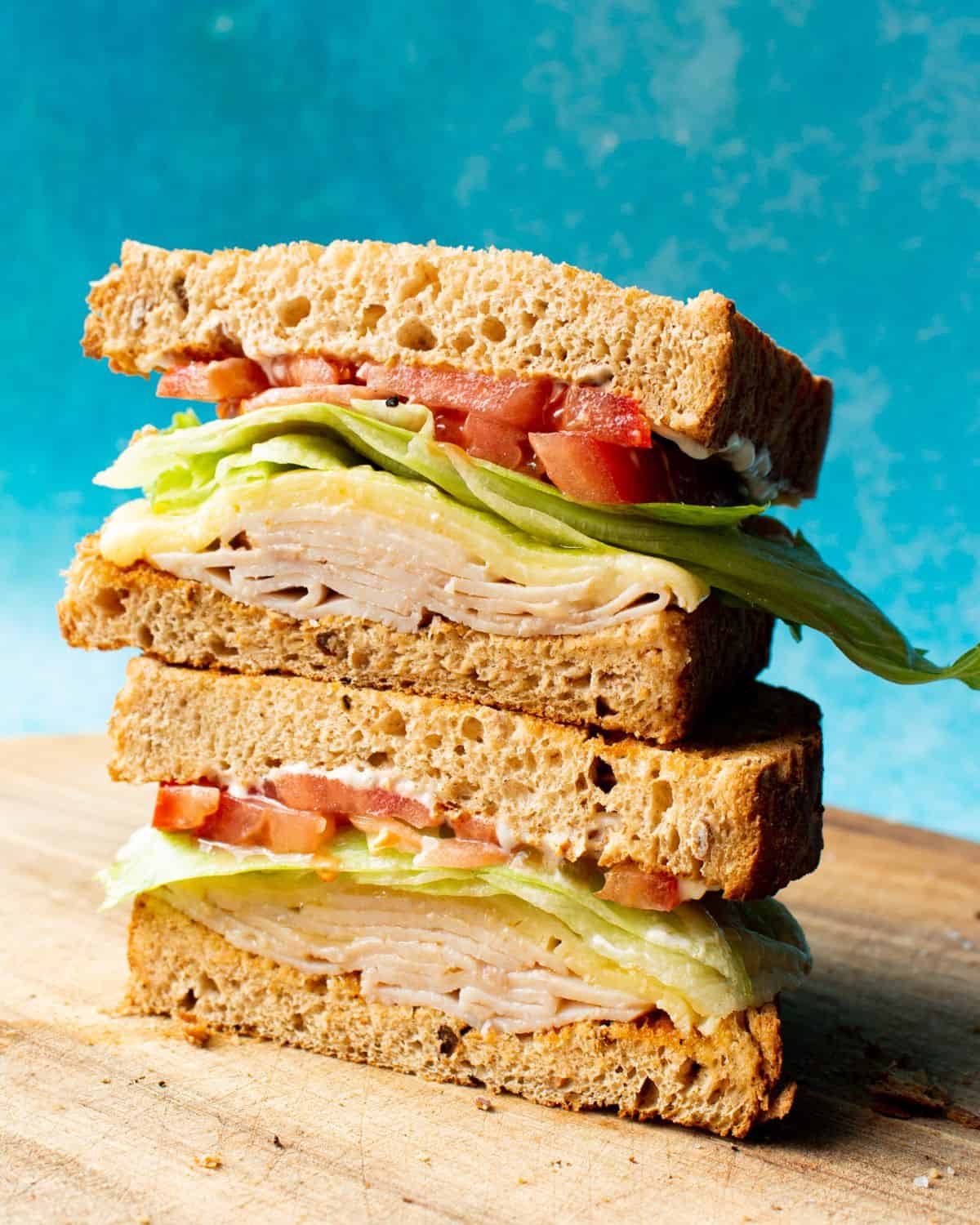Side view of huge turkey sandwich, with thick layers of tomato, lettuce, turkey and cheese piled on top of each on a wooden chopping board.