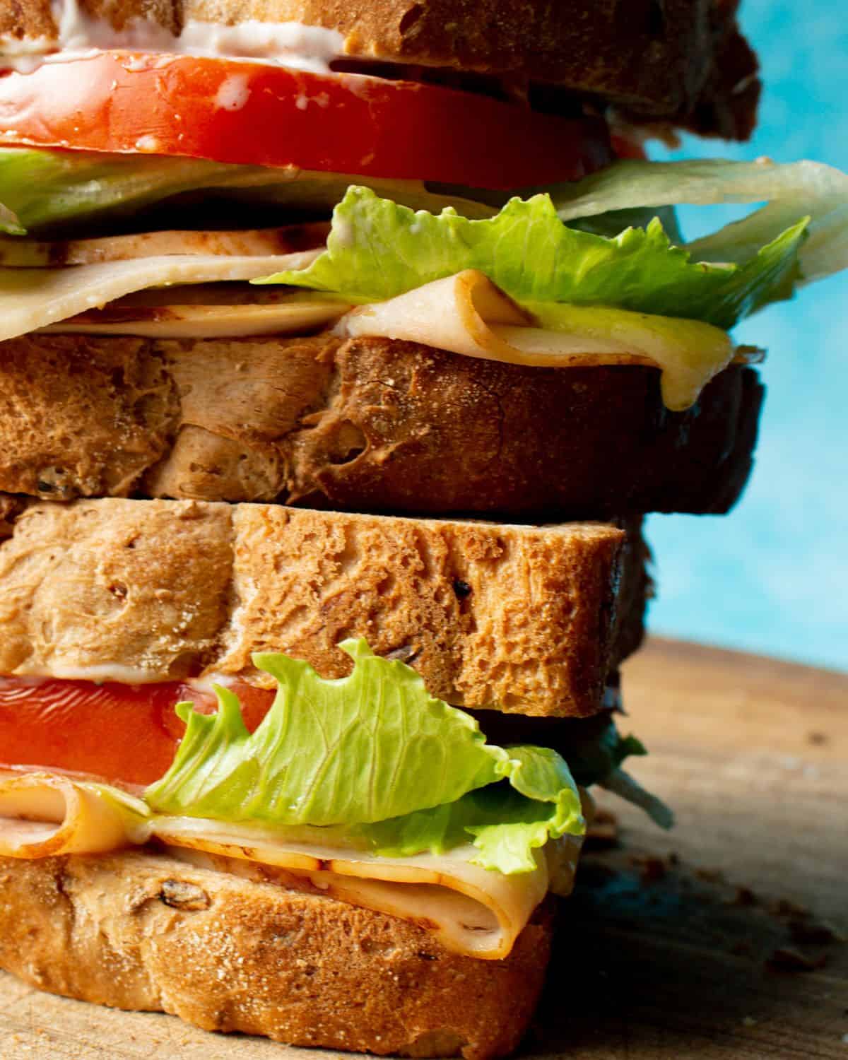 Close up view of turkey sandwich, with thick layers of tomato, lettuce, turkey and cheese piled on top of each on a wooden chopping board.