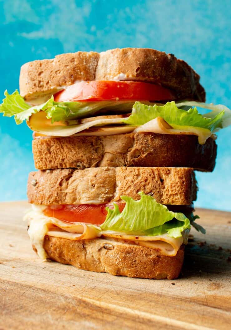 Side view of huge turkey sandwich, with tomato, lettuce and cheese piled on top of each on a wooden chopping board.