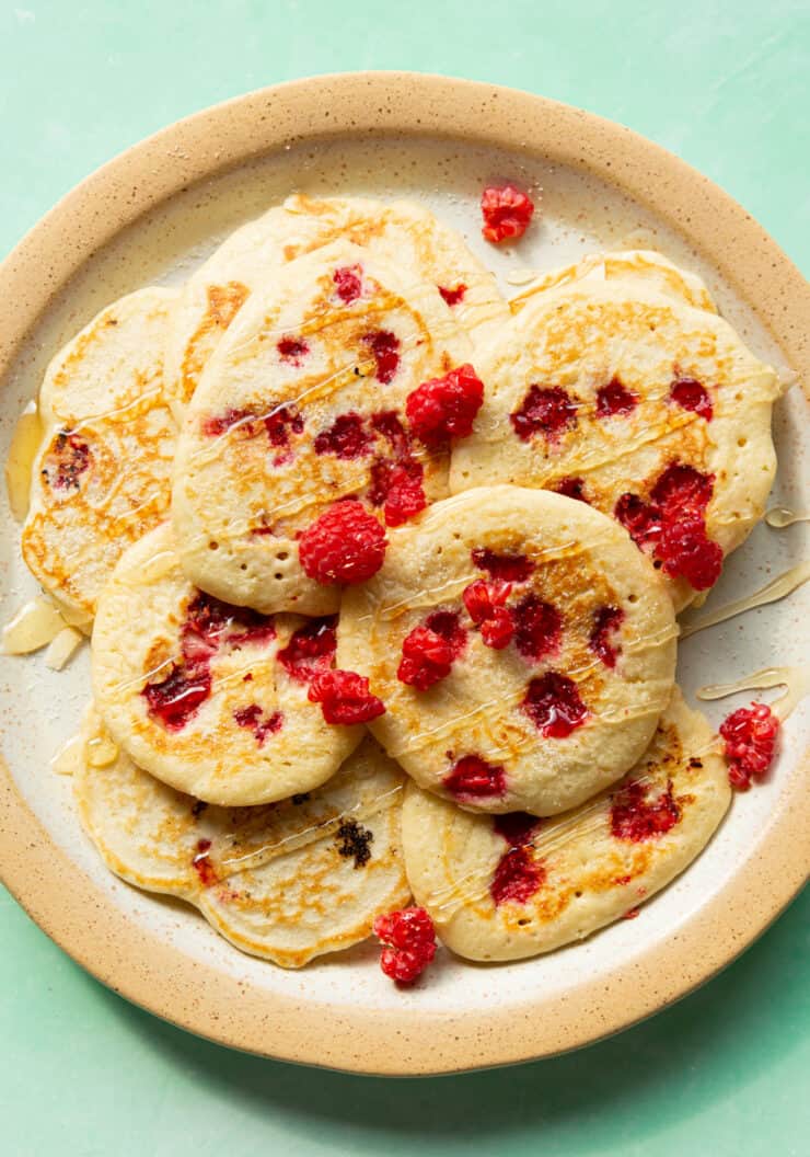 Lots of raspberry pancakes on a large plate and covered with a drizzle of honey and a few extra raspberries.