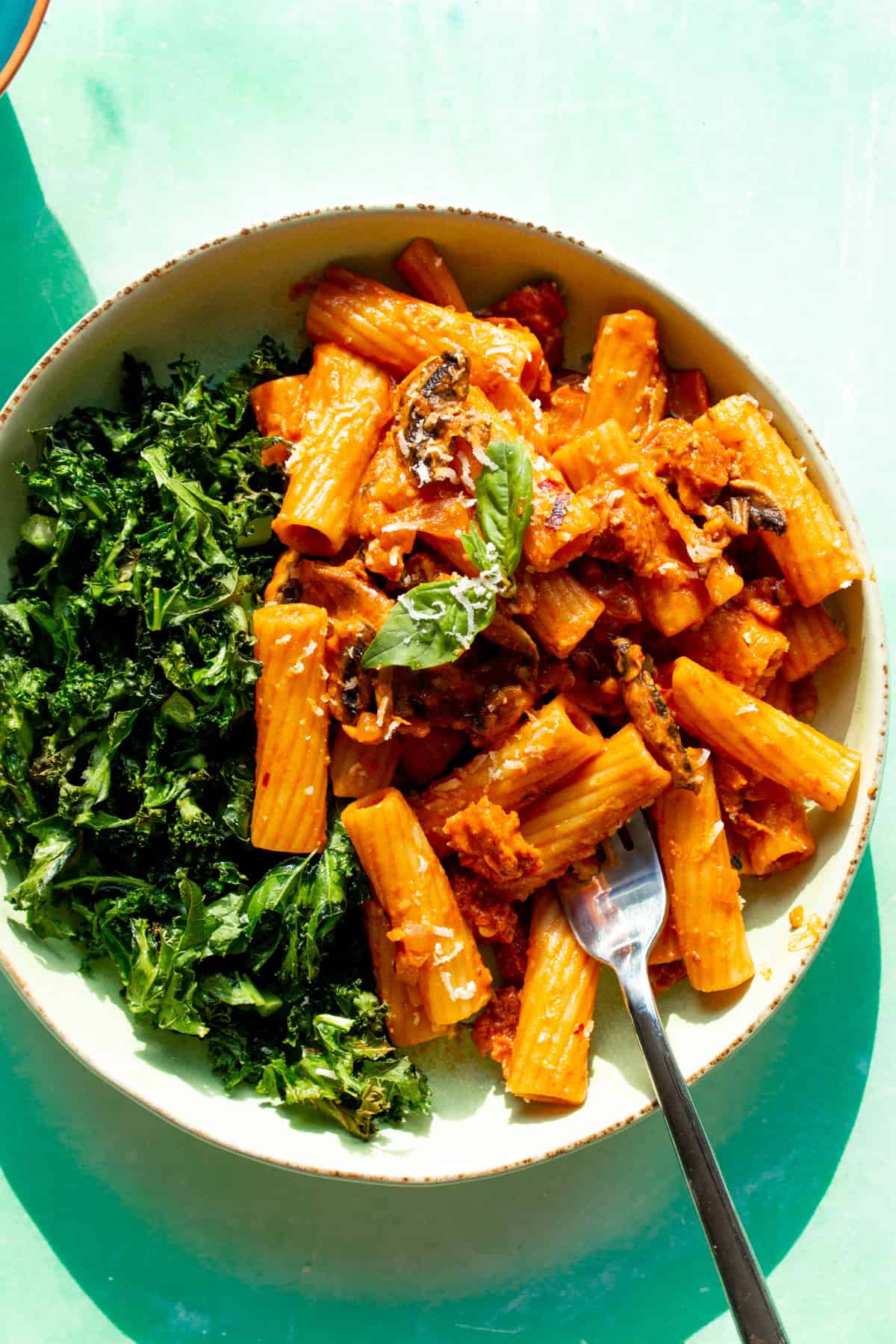 A bowl with rigatoni pasta with a tomatoey sauce with mushrooms, chorizo and fresh basil topping with some kale and a fork.