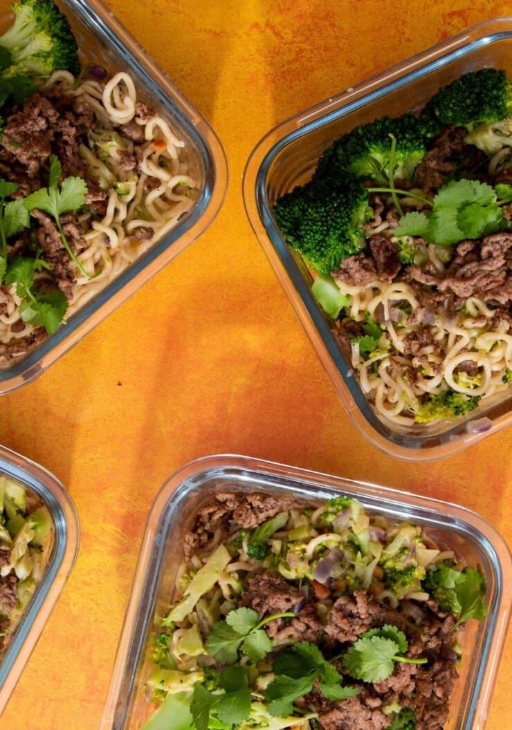 Broccoli & Beef Noodles topped with coriander and spring onion in 4 square glass meal prep containers on an orange background.