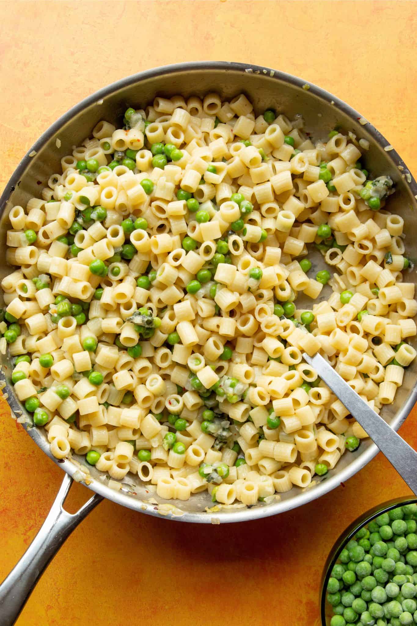 Pasta with peas with Ditali (short tubes of pasta) in a large stainless steel pan with metal spoon with bowl of peas.