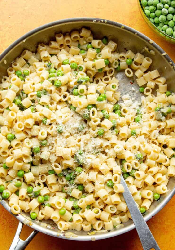 Pasta with peas with Ditali (short tubes of pasta), peas, topped with grated parmesan in a large stainless steel pan with metal spoon.
