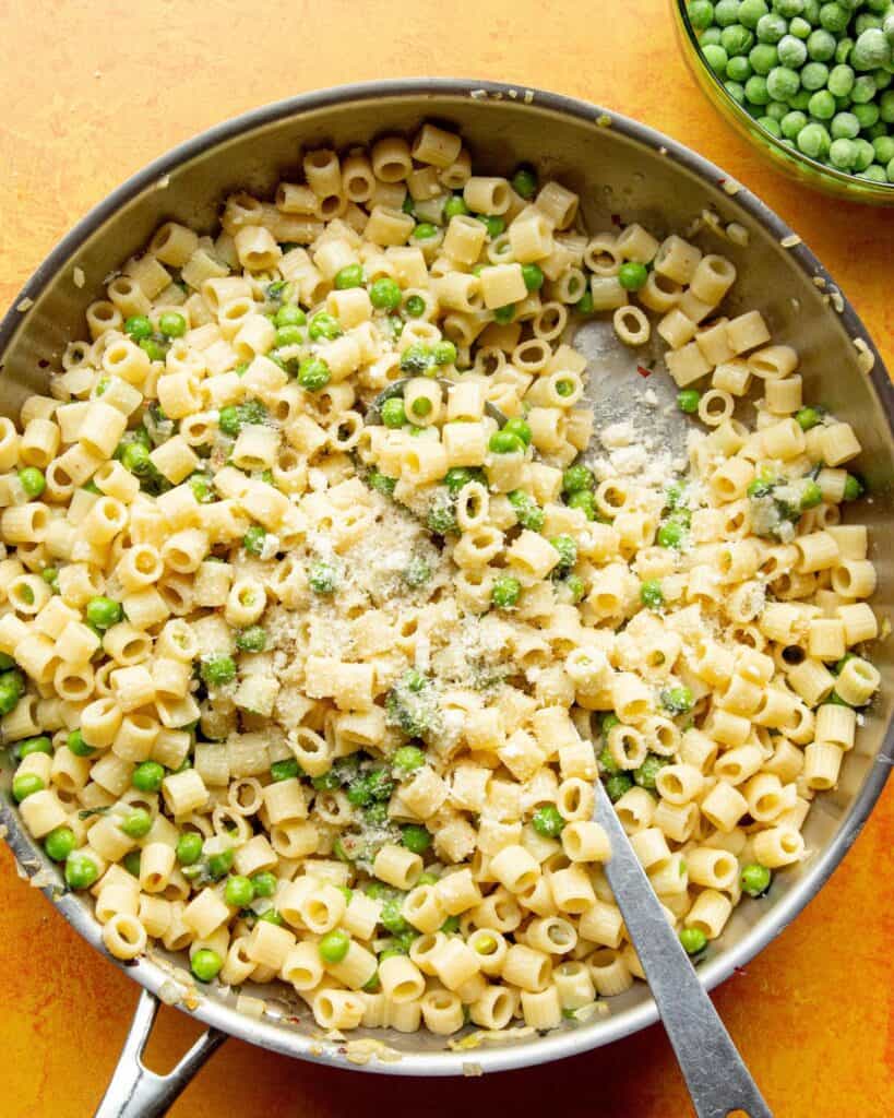 Pasta with peas with Ditali (short tubes of pasta), peas, topped with grated parmesan in a large stainless steel pan with metal spoon.