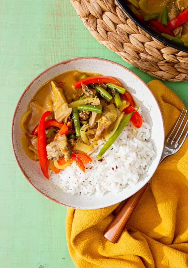 Chinese chicken curry with red peppers and green beans served in a bowl with rice, with a fork on a mustard cloth and a pan on straw mat.