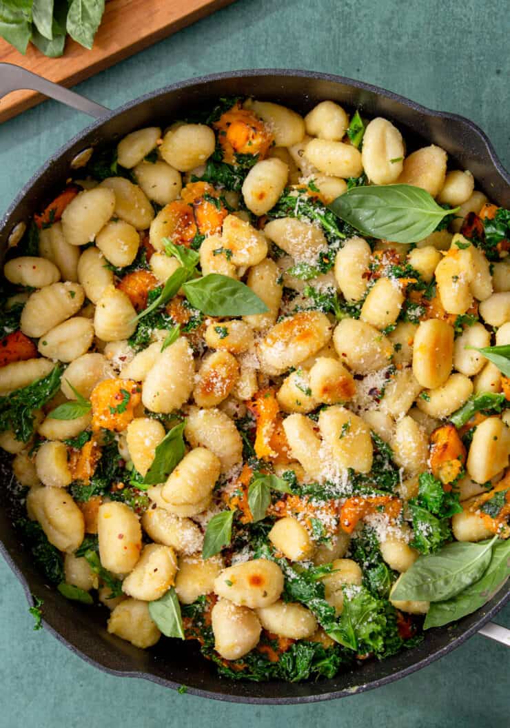 Golden browned and crispy pan-fried gnocchi with kale in a pan with a handle and garnished with parmesan and basil leaves.