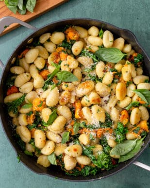 Golden browned and crispy pan-fried gnocchi with kale in a pan with a handle and garnished with parmesan and basil leaves.
