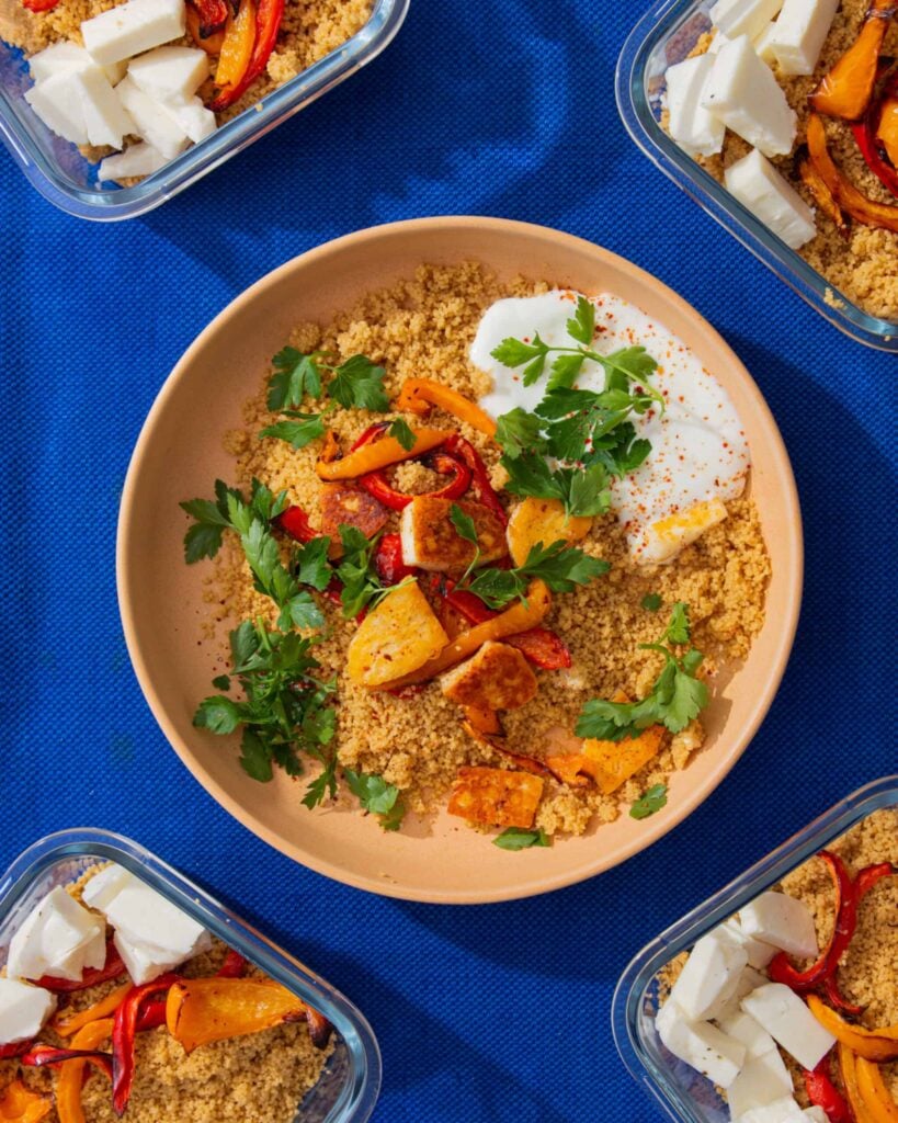 A bowl of spiced halloumi & couscous with peppers, yogurt and parsley and 4 meal prep containers with the halloumi, peppers and cous cous.