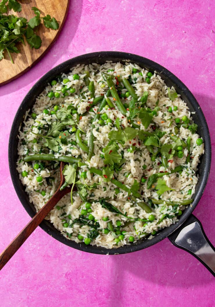 Coconut rice in a pan with green beans, peas and coriander with a wooden spoon on a pink background with coriander on a chopping board.