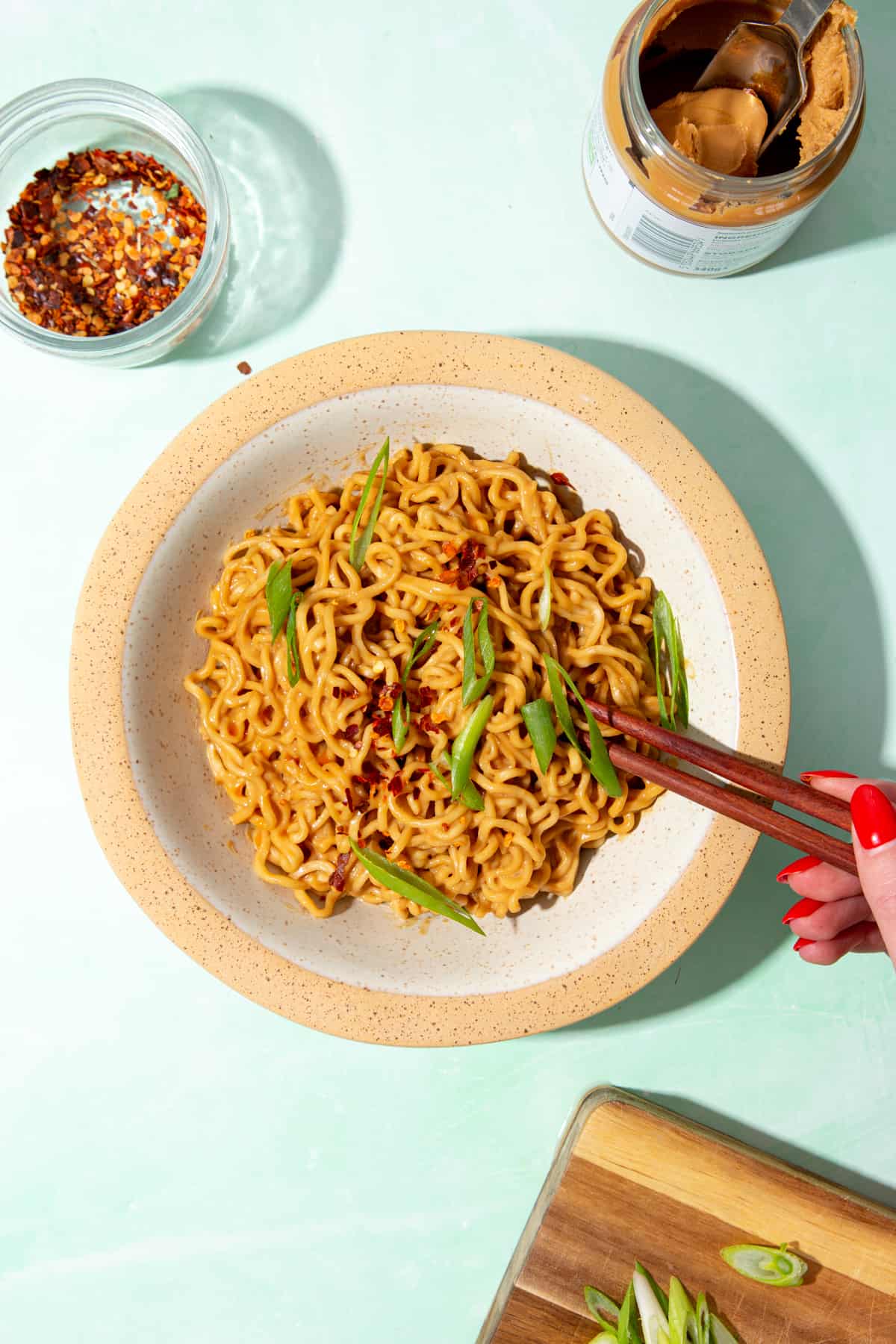 Noodles in a bowl with chop sticks and topped with slices of spring onion. Chilli flakes and peanut butter next to the bowl.