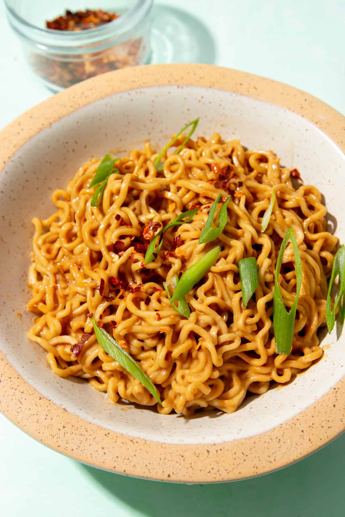 Noodles in bowl with slices of spring onion and chillies  and a glass bowl of chillies behind plate.