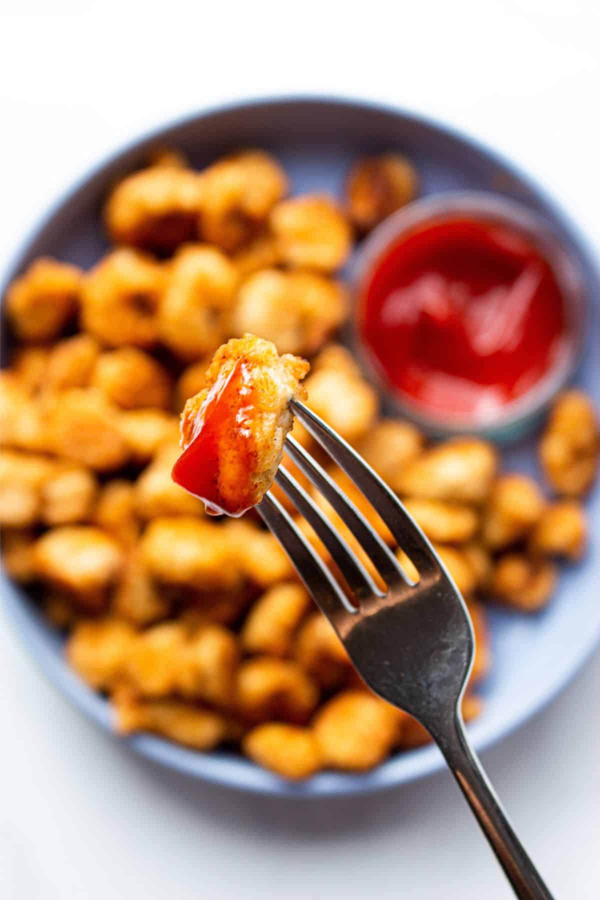 Close up of a pop corn chicken piece on fork with tomato ketchup over plateful of chicken pieces with tomato ketchup faded from view.