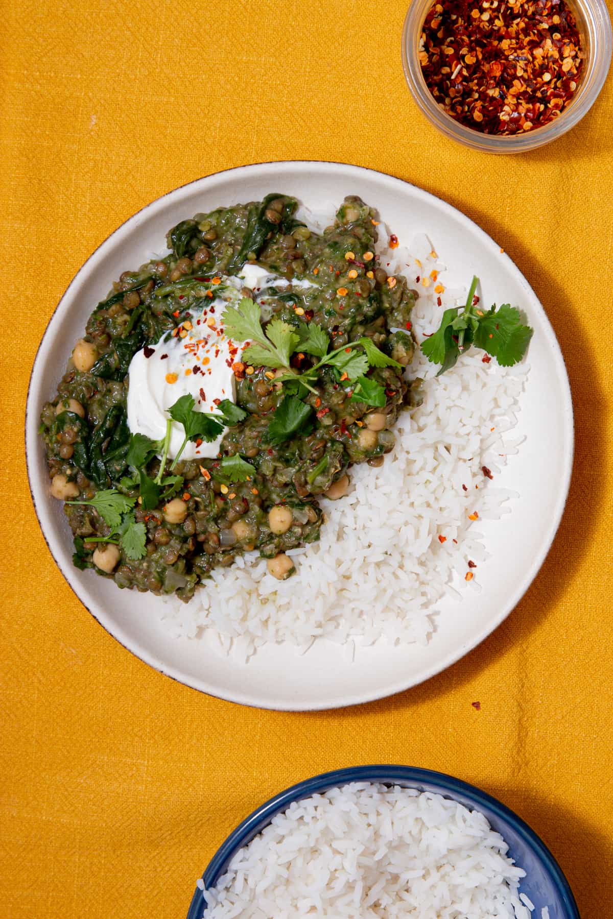 Lentil curry served on a plate with white rice and topped with yogurt and coriander, with chilli flake and extra rice in bowl.