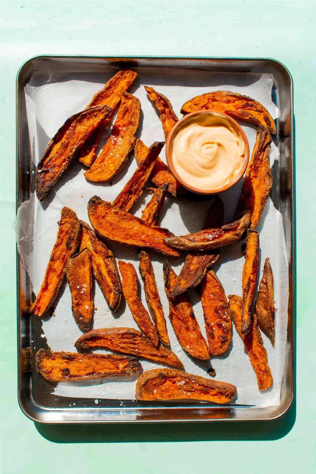 Crispy sweet potatoes laid out on parchment paper on a baking tray with dipping sauce.
