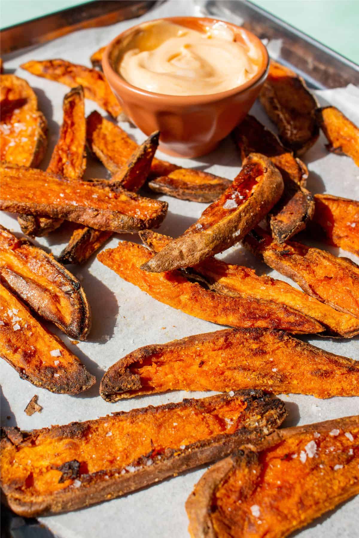 Side view of baked sweet potatoes fries on a baking tray with parchment paper and dipping sauce.