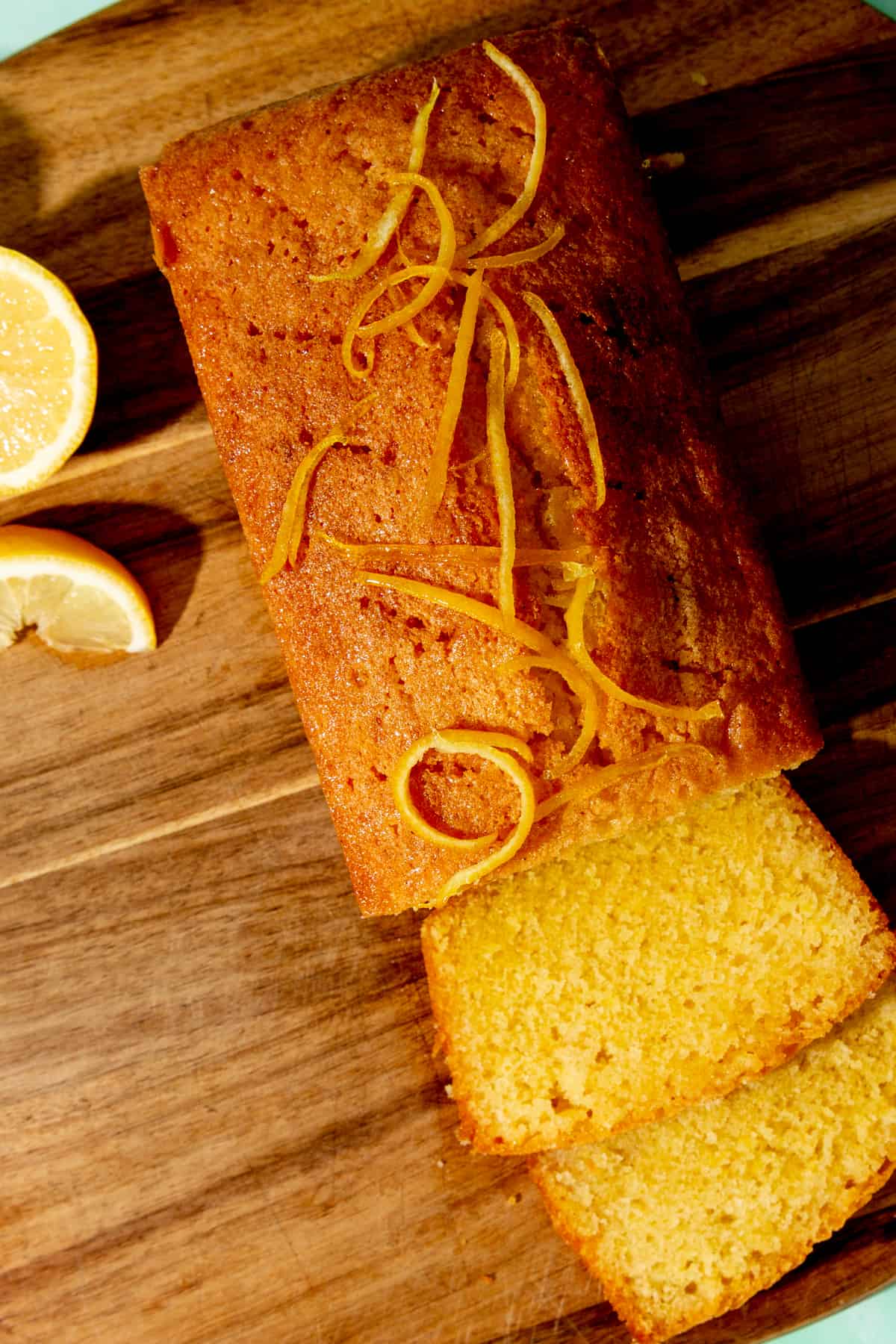 Overhead shot of golden browned rectangular cake with a slice cut in front and lemon sliced on the wood board background.
