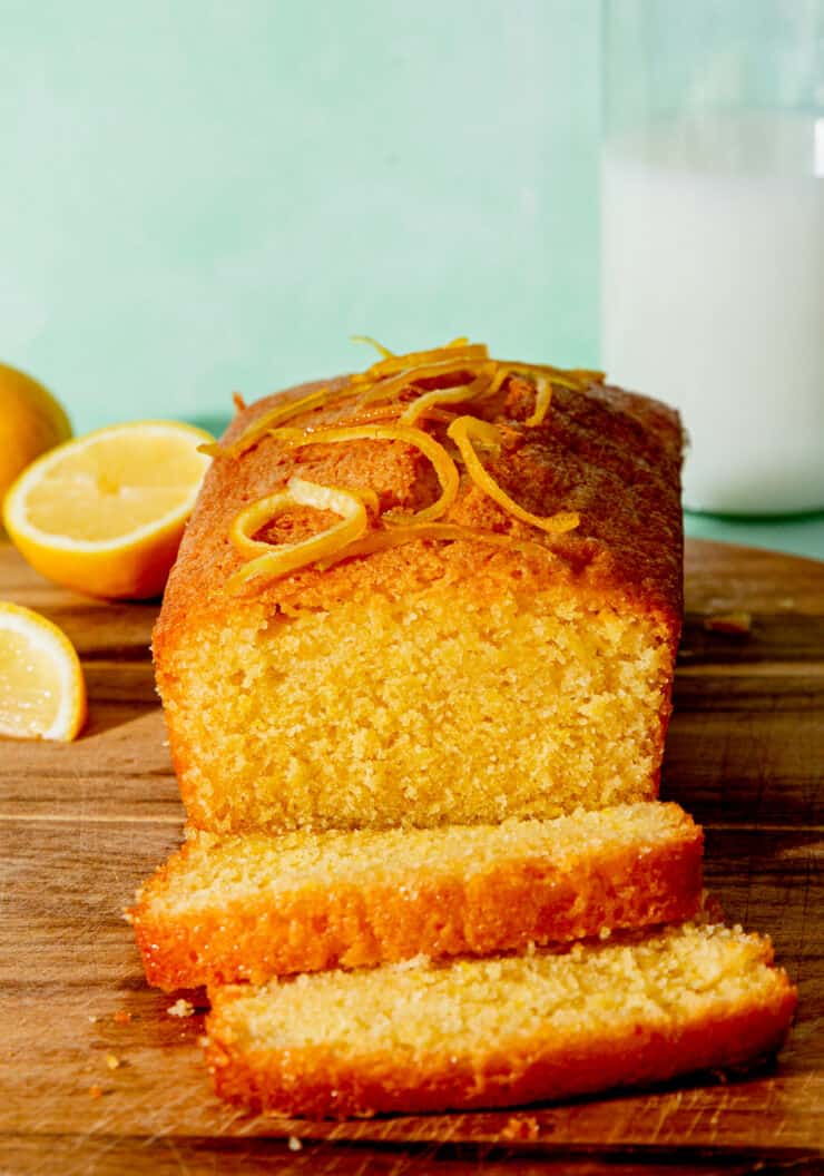 A cake sliced with lemon rind topping, lemon wedged and milk in the background.