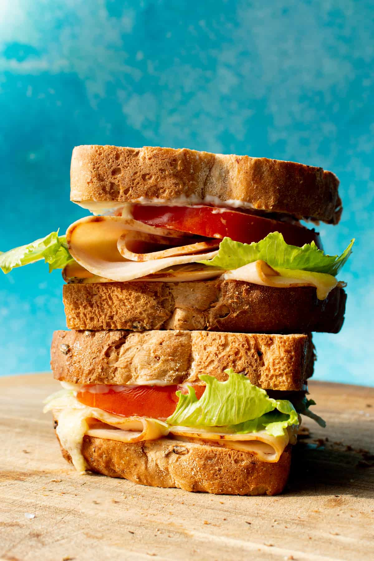Side view of huge turkey, tomato and lettuce sandwich with thick slices of bread and a blue background.