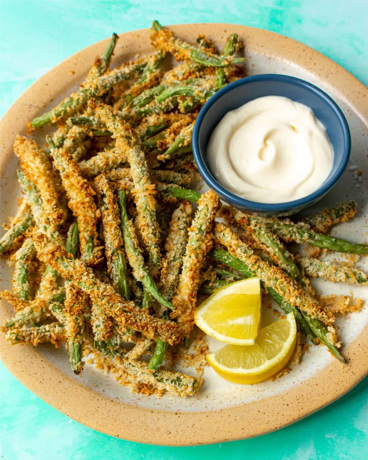 browned and battered green beans on plate with dipping sauce and lemon wedges.