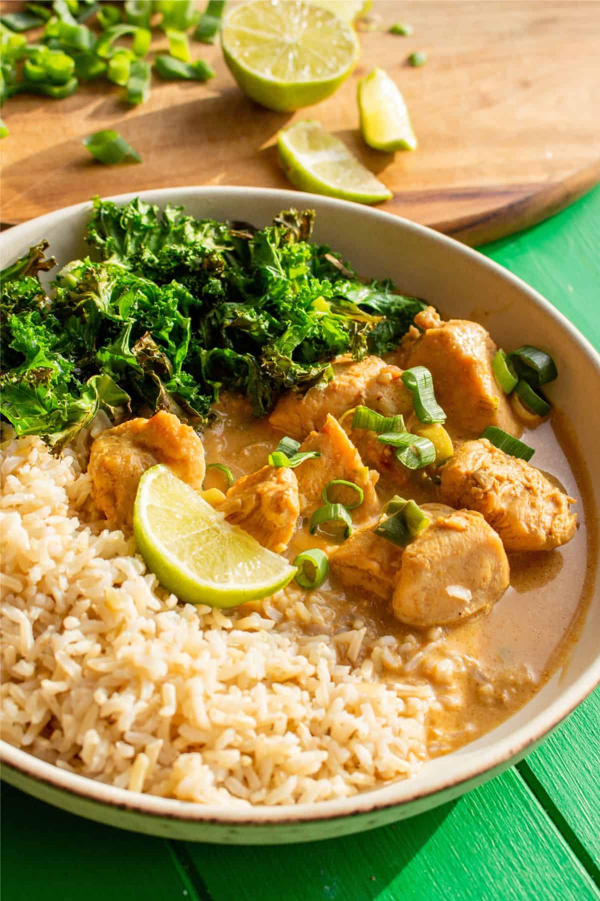 Chicken Satay Curry with brown rice, a wedge of lime and kale next to a wooden chopping board with lime wedges and spring onion.