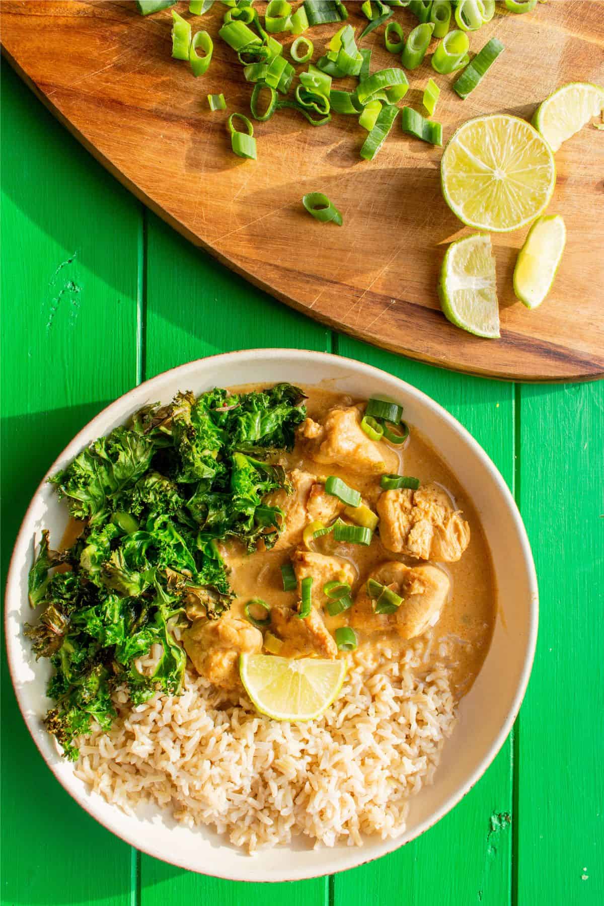 A bowl of Chicken Satay Curry with brown rice and kale with lime wedges and spring onion slices on a chopping board.