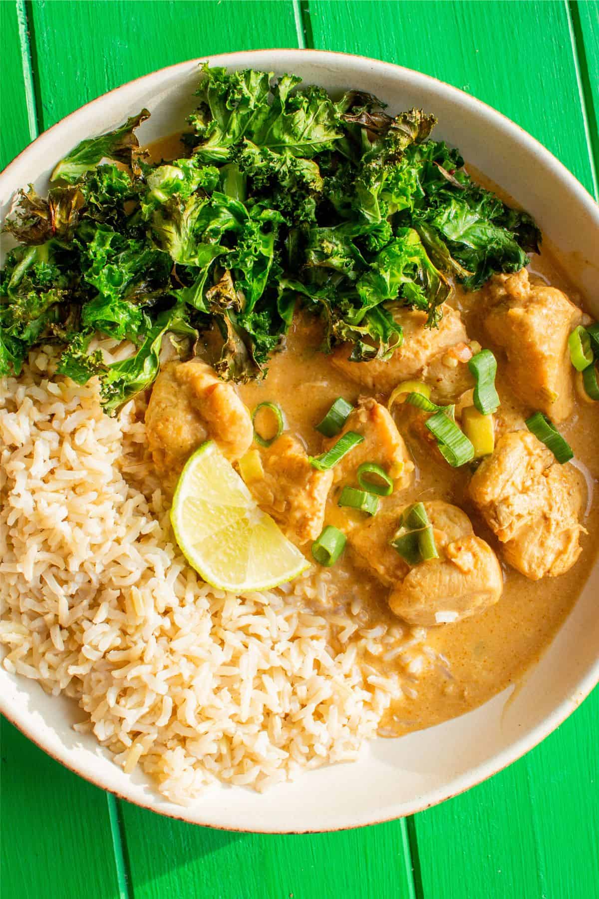 A bowl of peanut butter chicken curry with brown rice, kale and a wedge of lime on a green background.