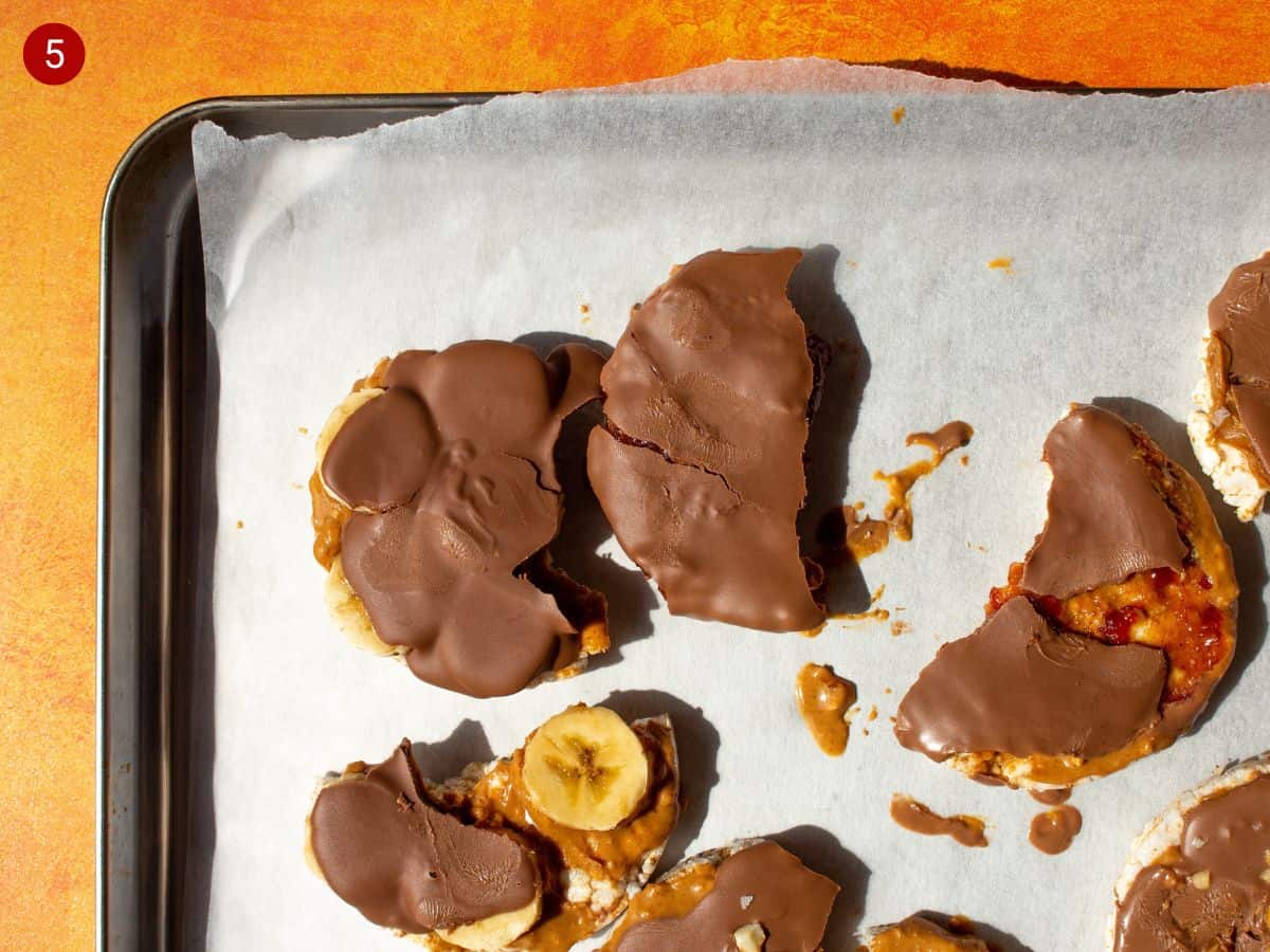 Rice cakes topped with peanut butter and chocolate broken in pieces on parchment paper on tray.
