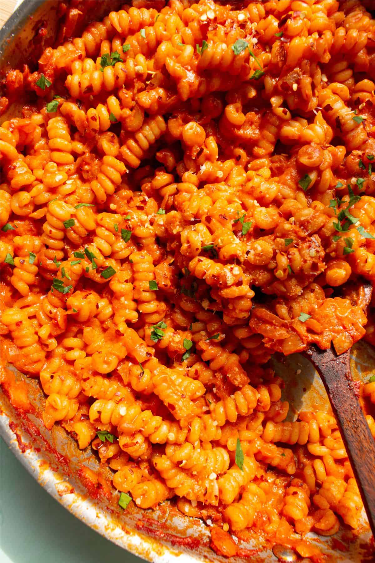 Close up of creamy tomatoey fusilli pasta (garnished with chopped parsley) in pan with wooden spoon.