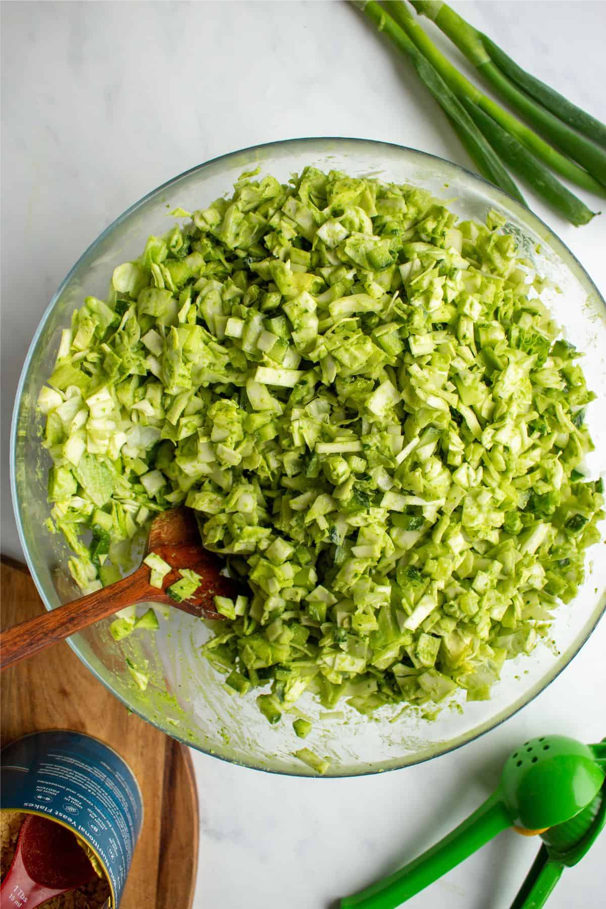 Overhead shot of large glass bowl of chopped salad with green dressing mixed with a wooden spoon and spring onions and nutritional yeast in background.