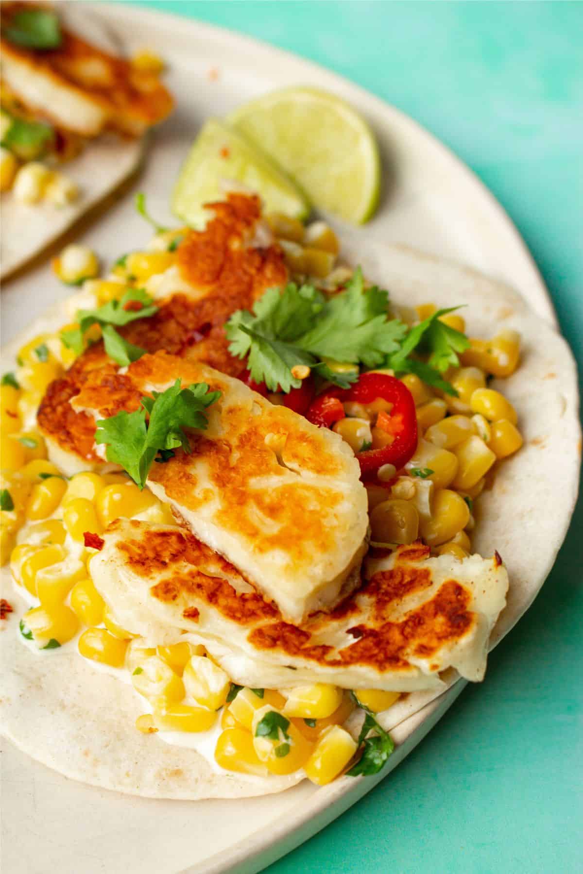 Close up photo of a white tortilla with sweet corn, golden browned halloumi, chillies, coriander and lime wedges.