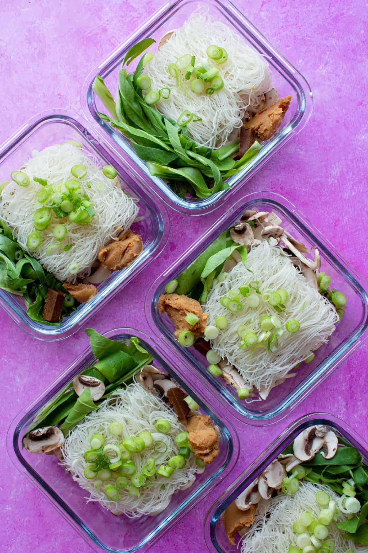 Healthy instant noodles ingredients in 5 glass meal prep containers, with dry noodles, miso paste, slices of spring onion and pok choi.