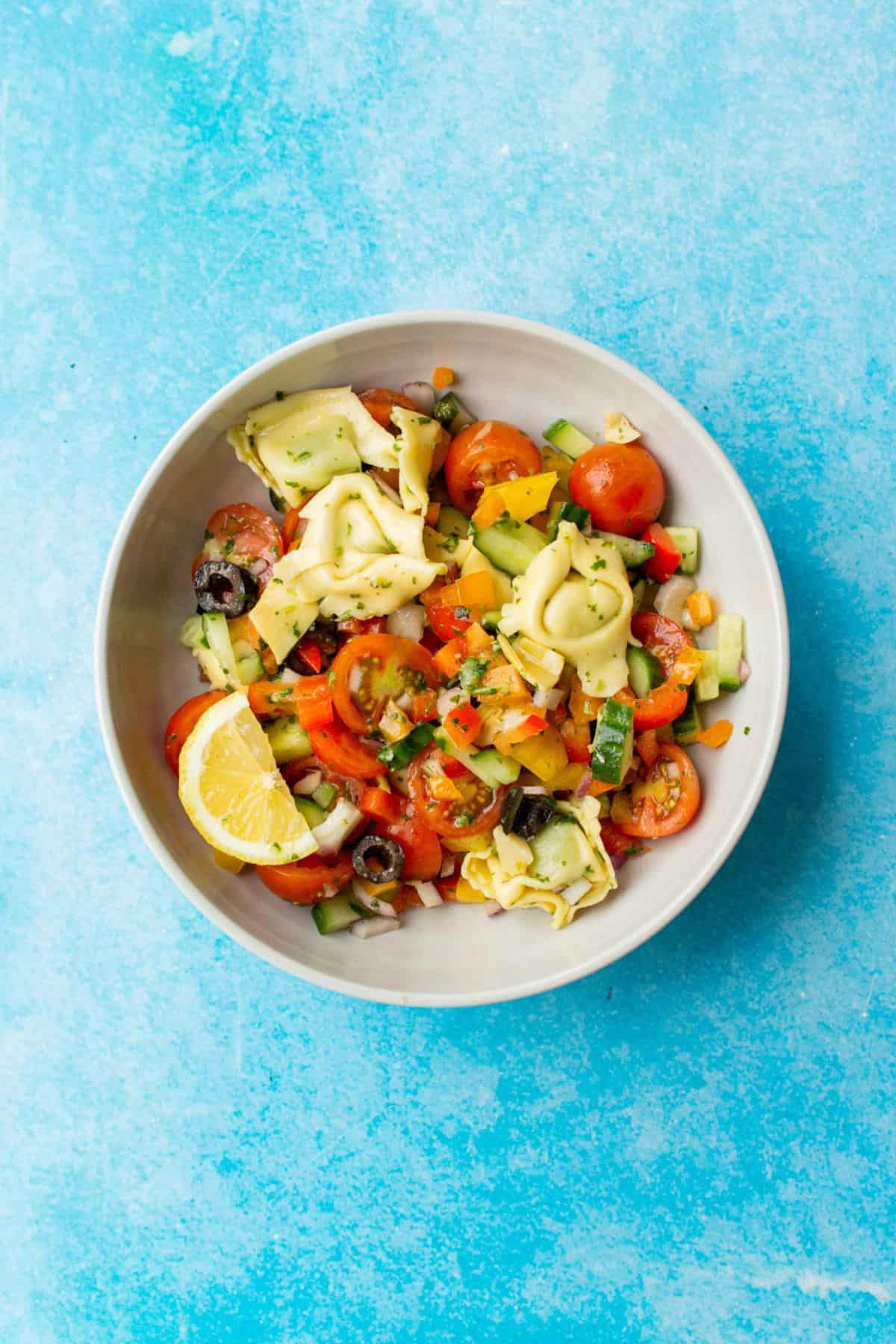 Pasta salad with chopped vegetables with a lemon wedge in a white bowl on a blue background.