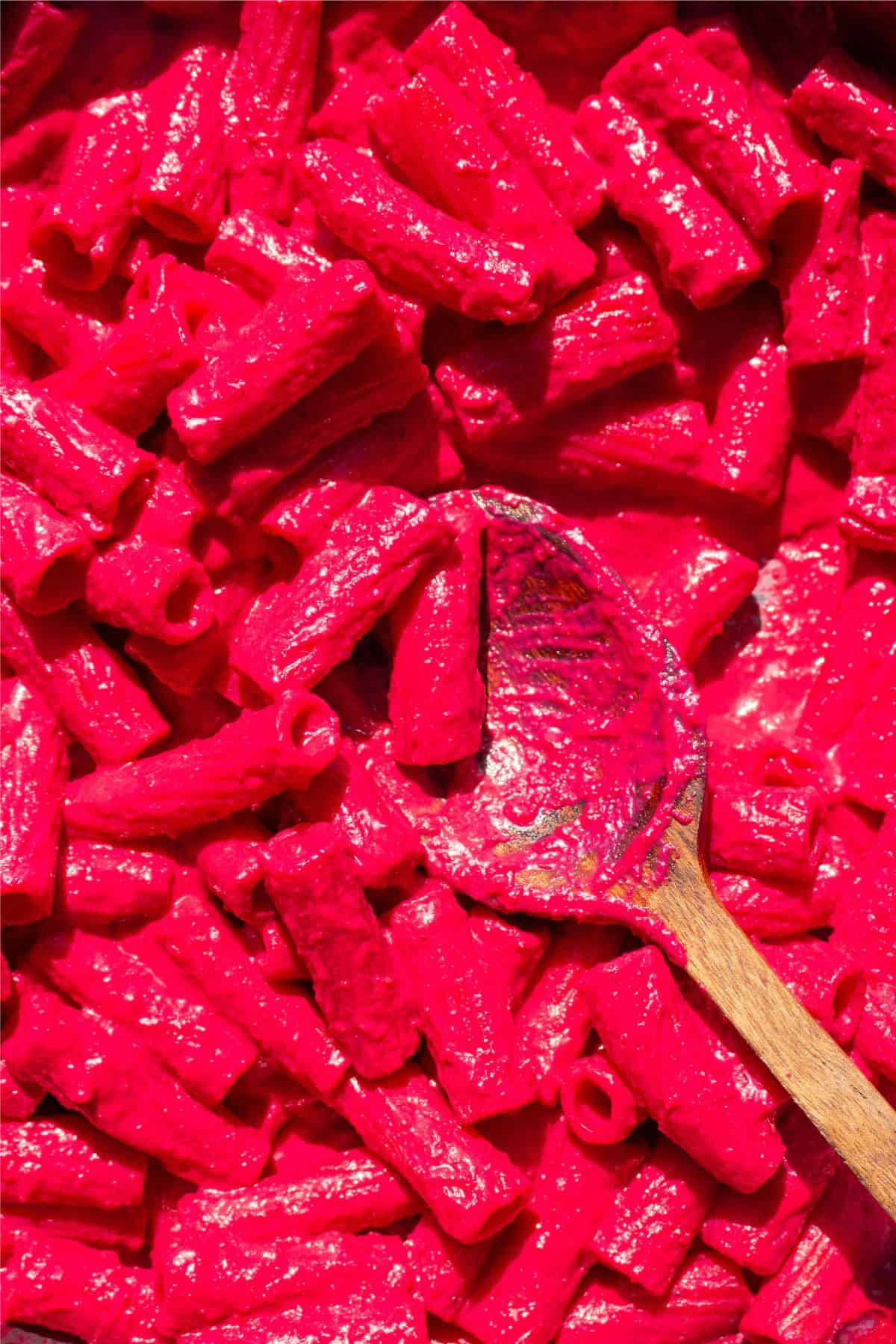 Close up of pasta with rigatoni covered in pink beetroot sauce mixed with wooden spoon.