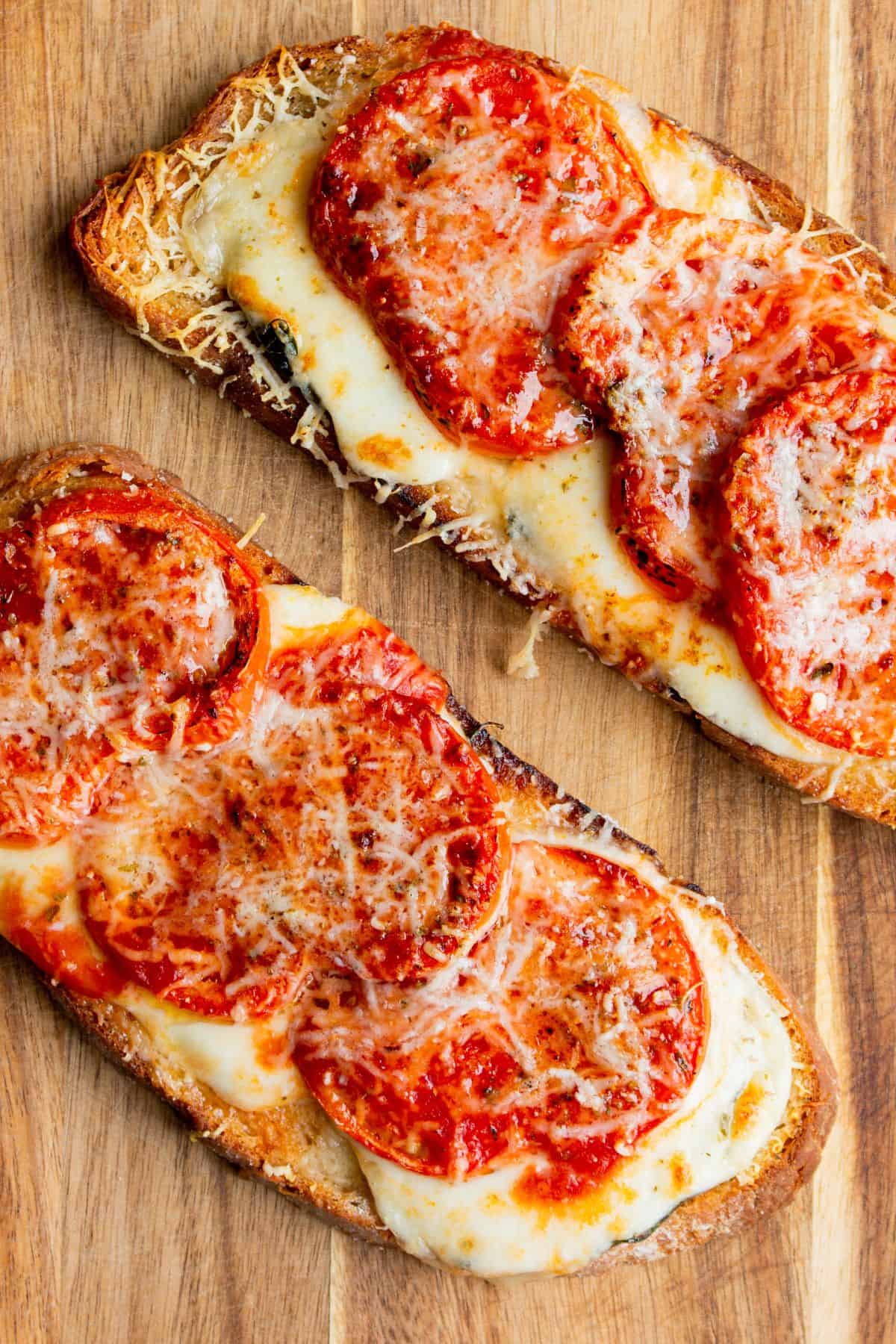 Close up of 2 pieces of pizza toast with golden browned melted cheese and roasted tomatoes on a wooden board.