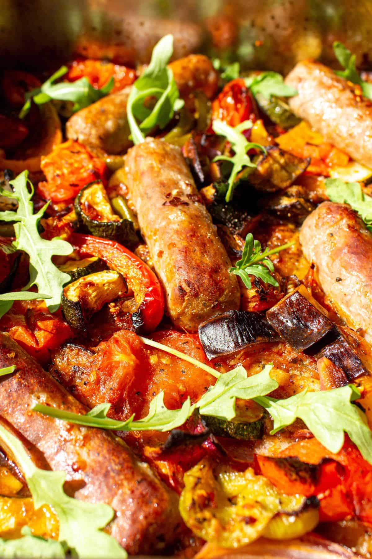 Close up of cooked tray bake with roasted vegetables and sausages garnished with rocket.