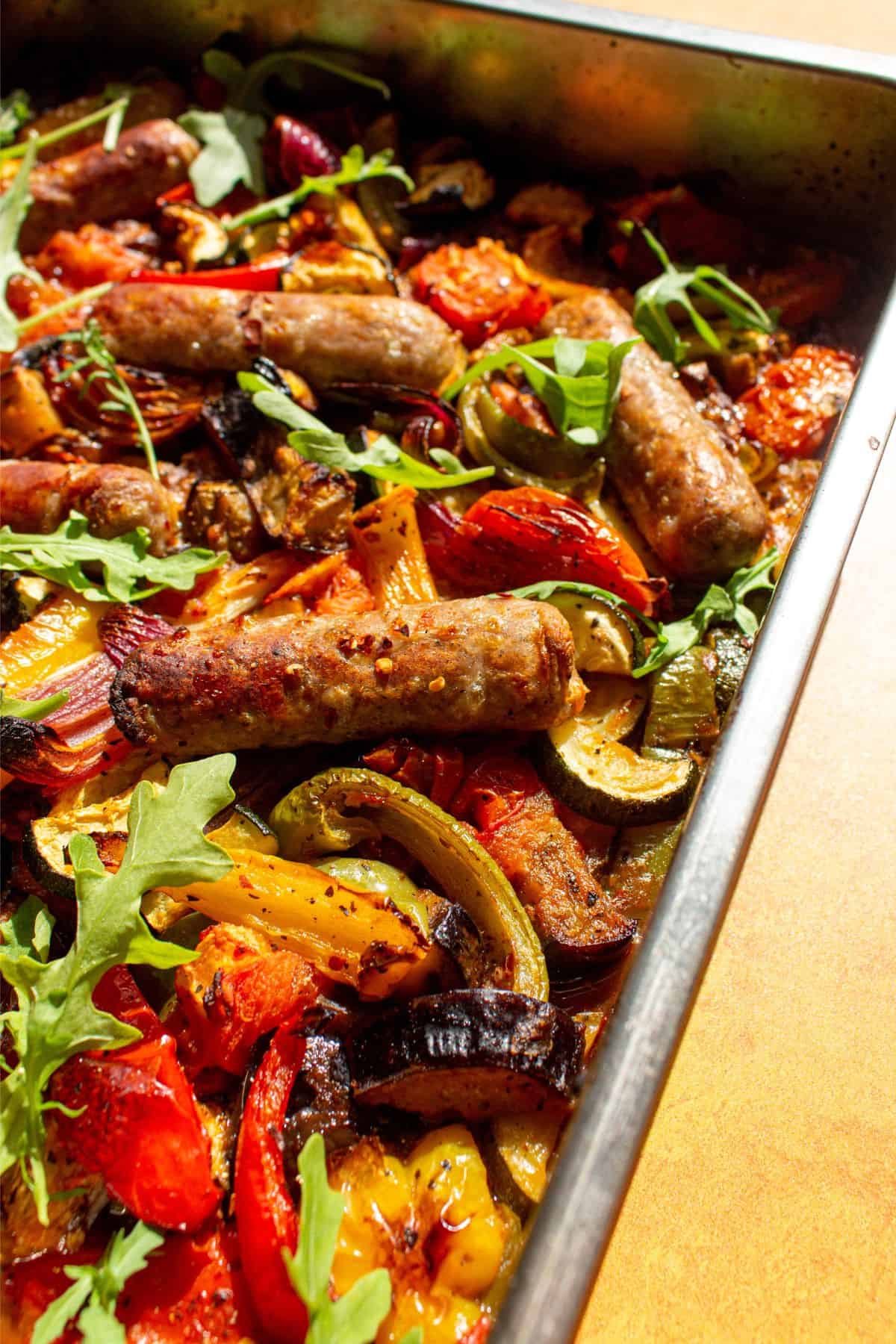 Close up of roasted sausages and peppers, courgettes, red onion and courgettes) in stainless steel baking tray on a yellow background.