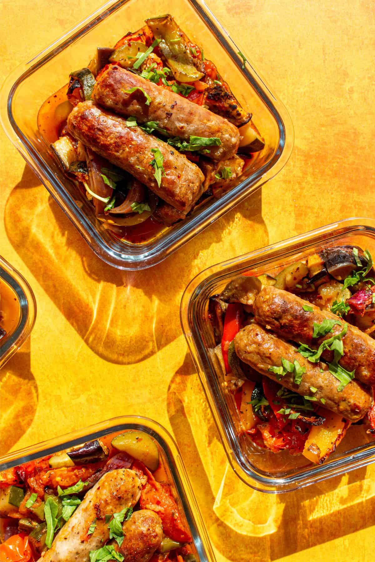 Roasted vegetables and 2 sausage in 4 separate, square, glass meal prep containers on a yellow background.