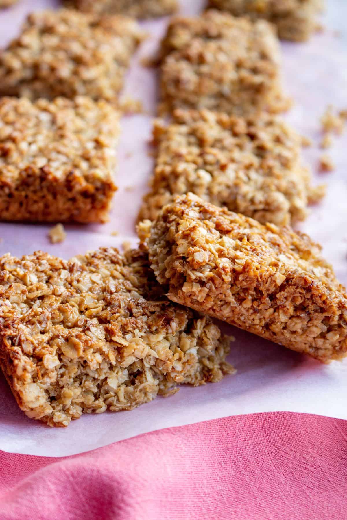 Flapjacks cut in rectangular pieces and scattered over parchment paper with crumbs on a pink cloth.
