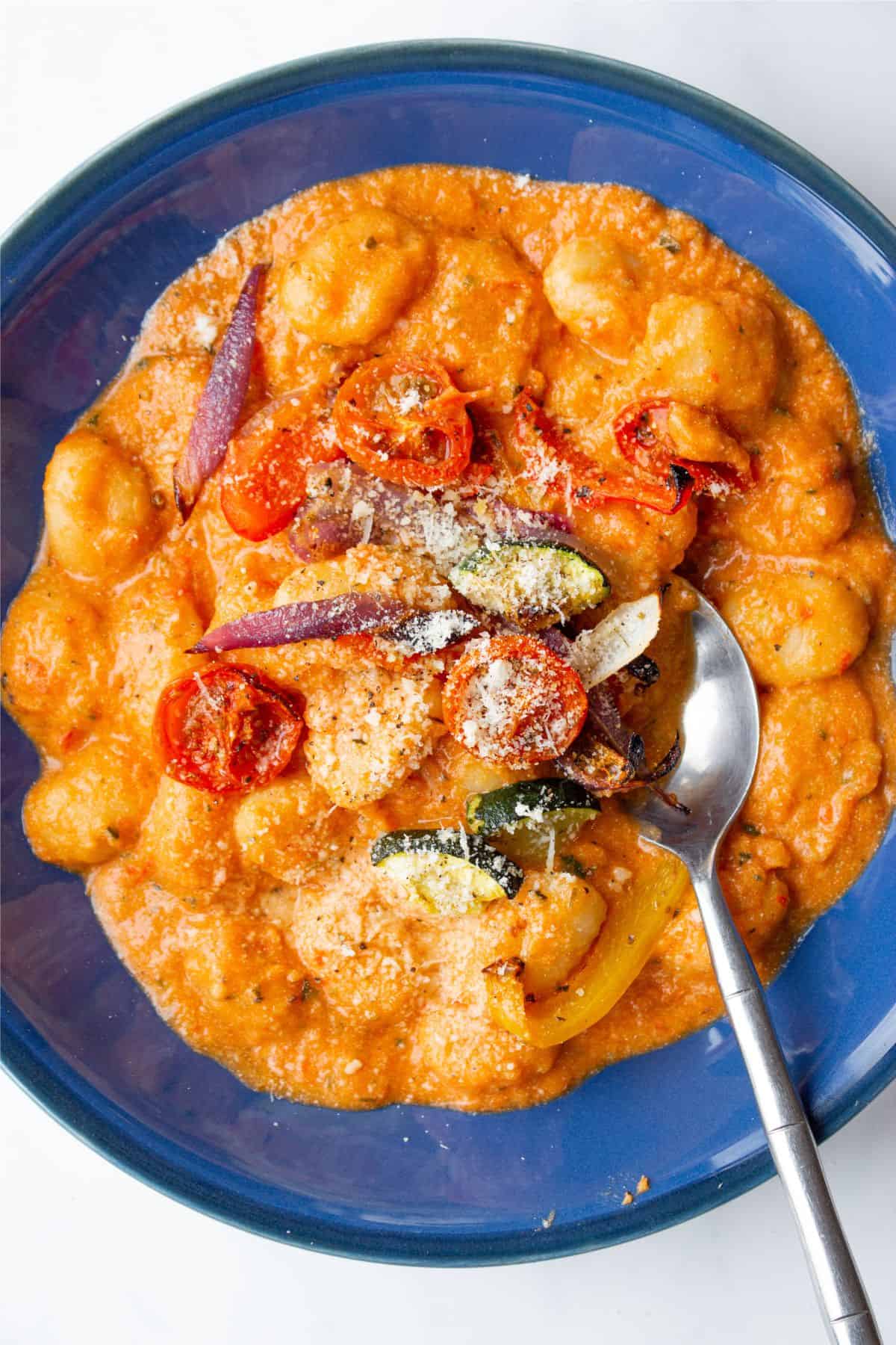A bowl of gnocchi with creamy tomato sauce, roasted vegetables and parmesan with a metal spoon in a blue bowl.