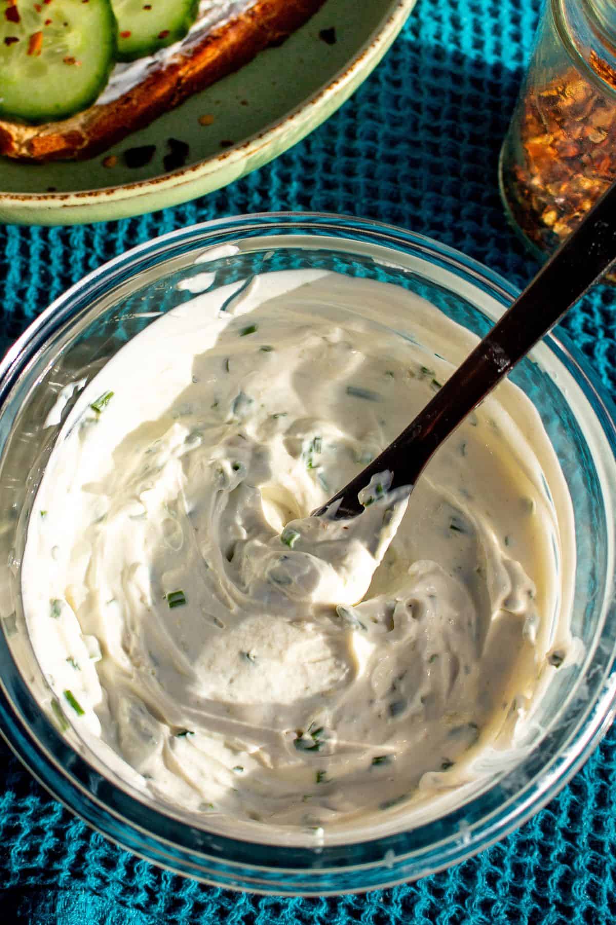 Garlic and herb cream cheese in a glass bowl on a blue cloth with chilli flakes in background and plate with cucumber on cream cheese toast.