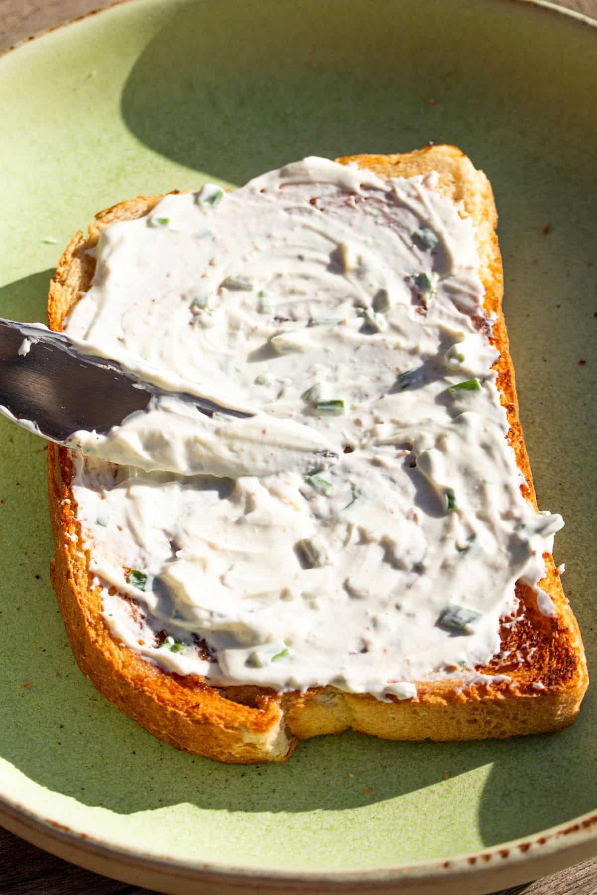 A knife used to spread garlic and herb cream cheese onto piece of toast in a blue bowl.