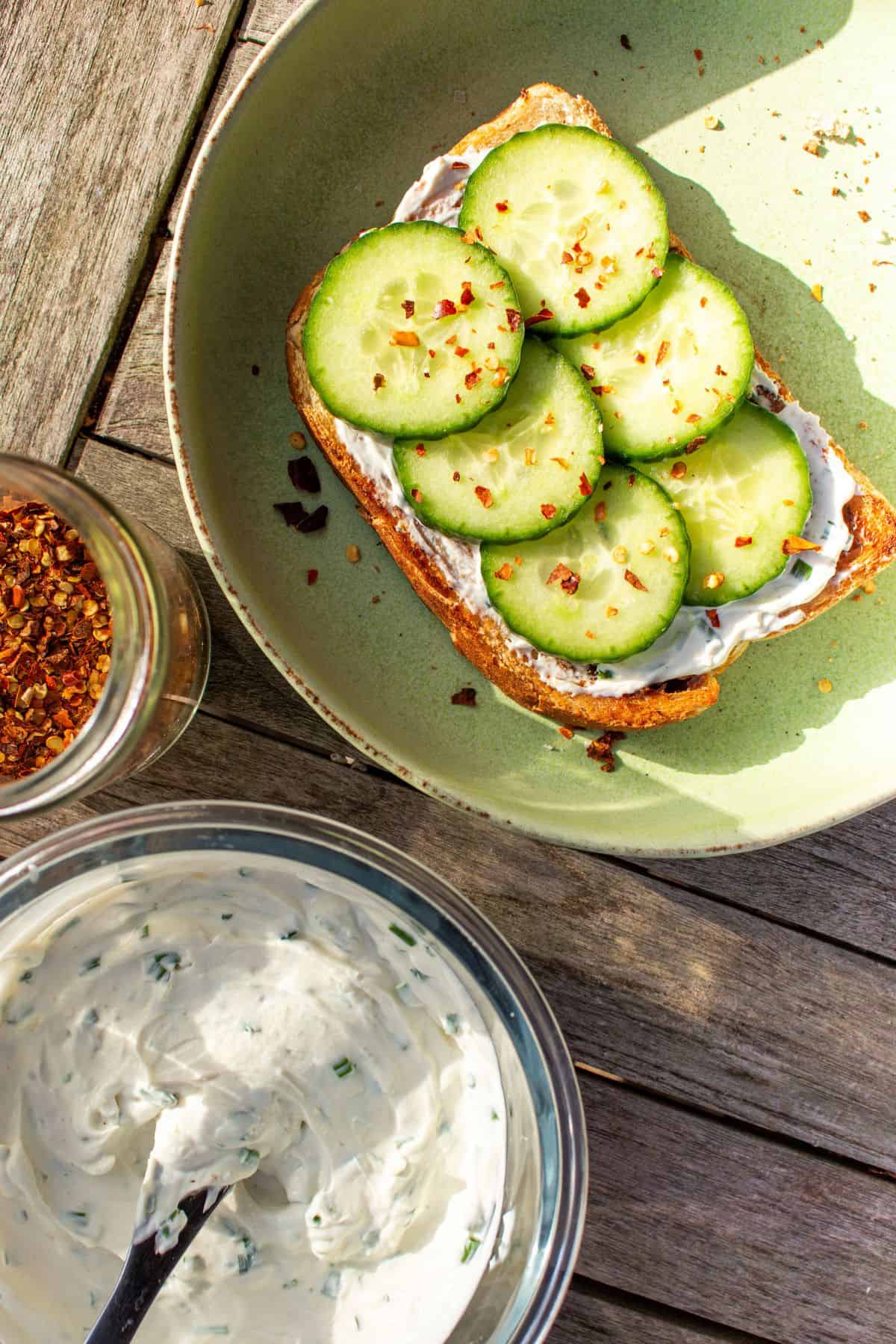 A pale green plate with 1 slice of toast layered with cream cheese and cucumber slices, a bowl of cream cheese and chilli flakes.