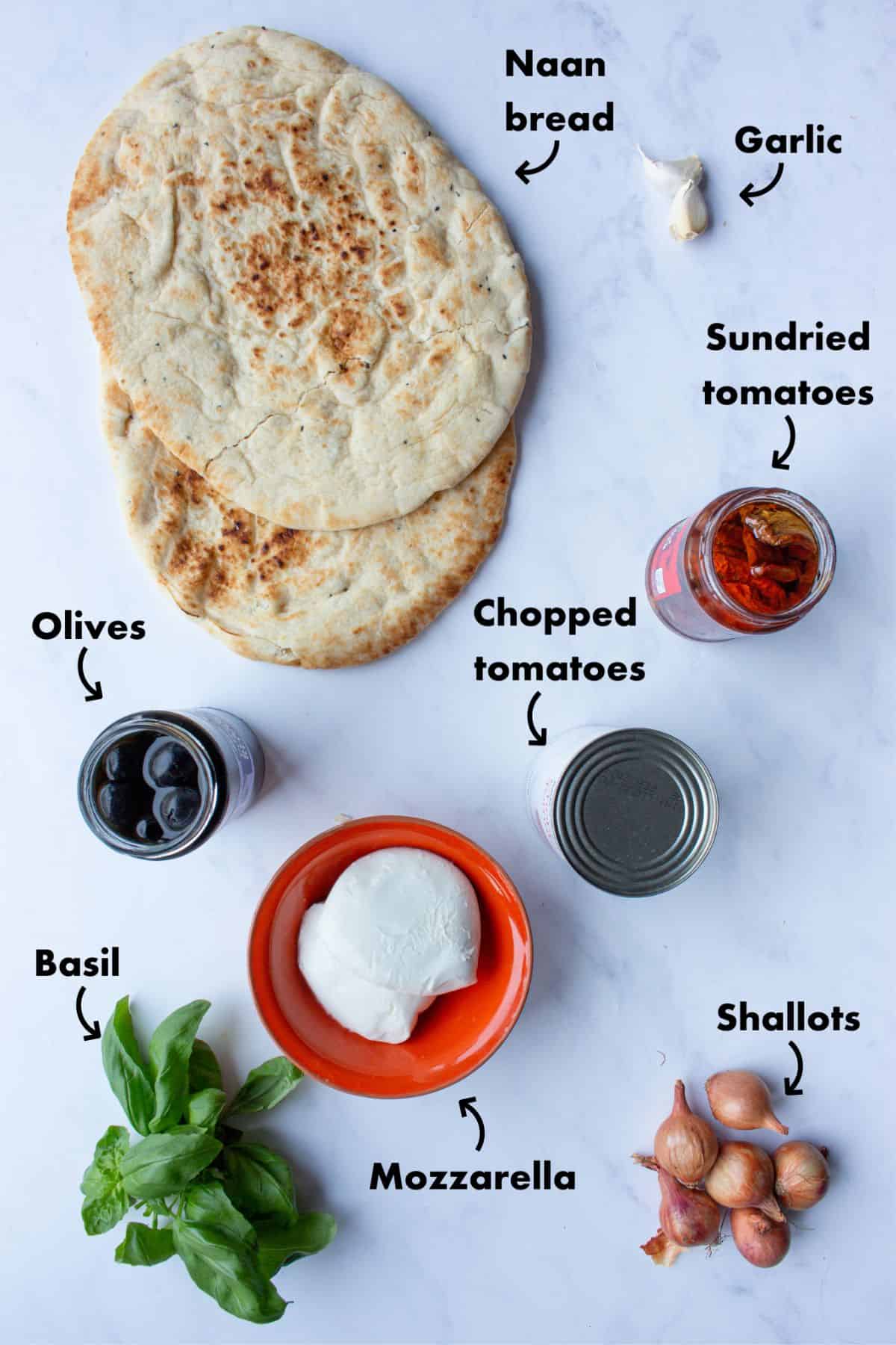 Ingredients to make naan bread pizzas laid out on a greyish background and labelled.