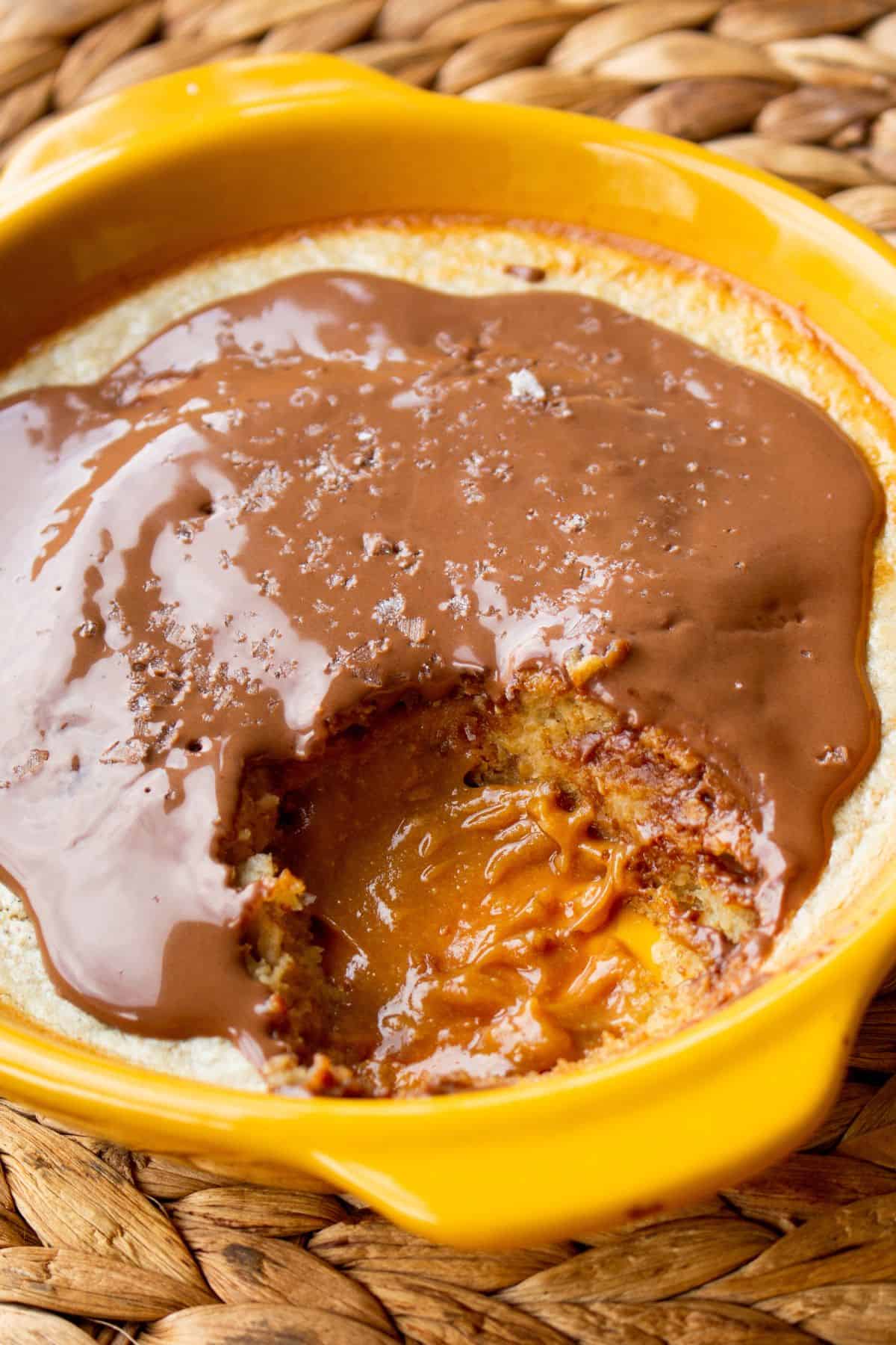 A yellow ramekin with the baked oats with a melted chocolate topping with a piece taken showing the gooey peanut centre on a straw heat mat.