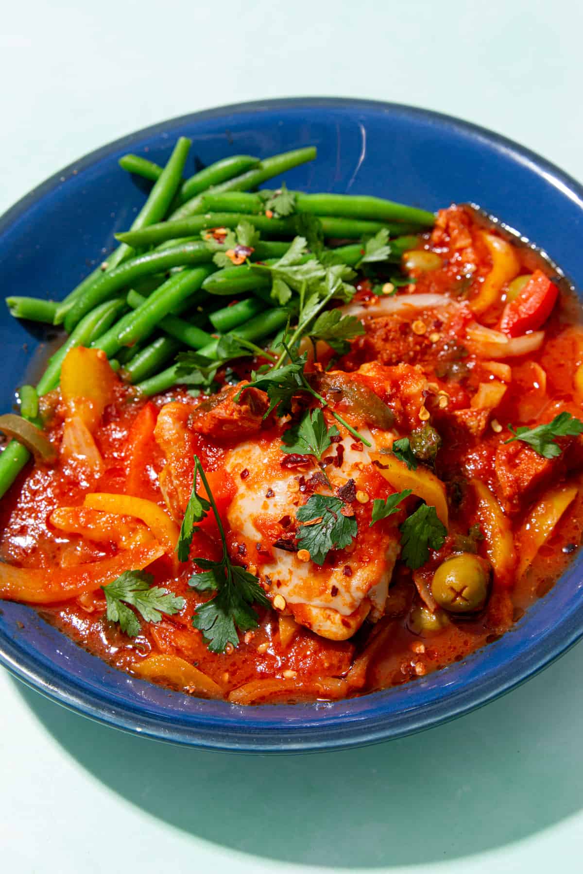 Chicken in a tomato sauce with green olives and green beans on a blue plate.