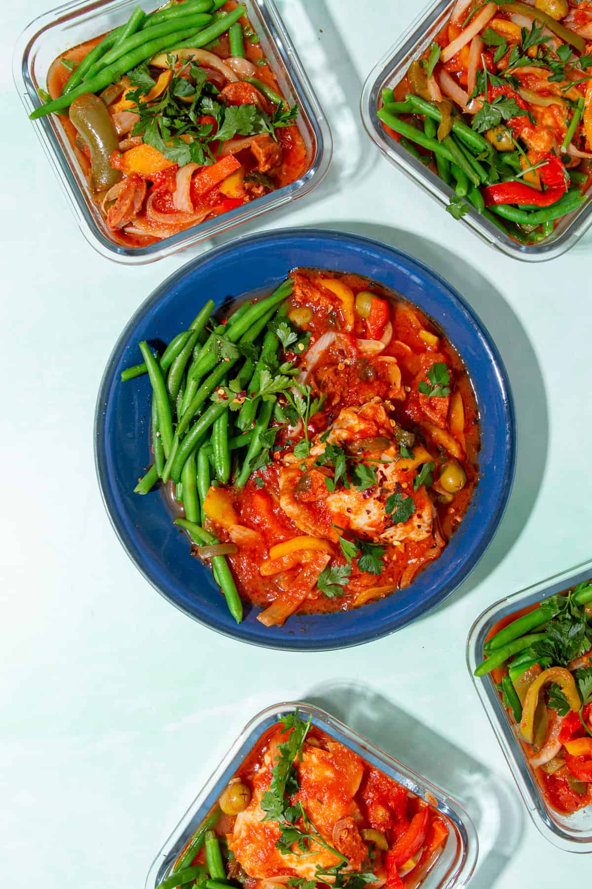 A blue dish with chicken in a tomatoes sauce with olives and peppers and a side of green beans, with 4 square glass meal prep containers filled with the same ingredients.