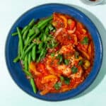 A blue bowl of chicken and chorizo in a tomatoes sauce with peppers, a side of green beans and some chilli flakes in a small bowl at side.