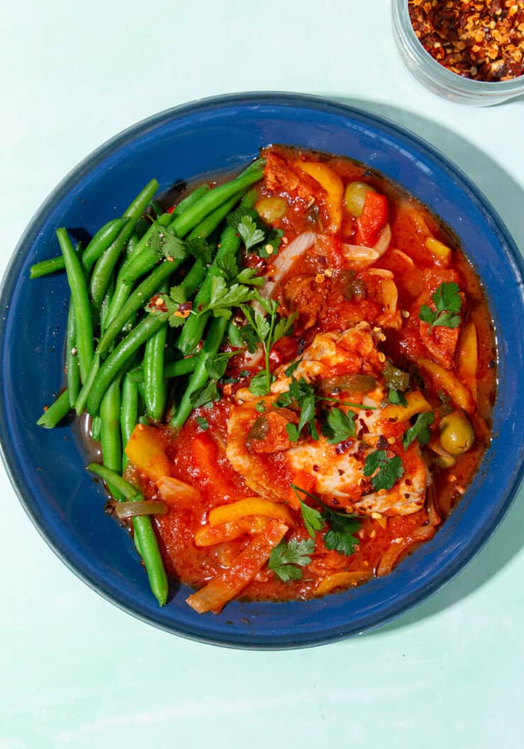 A blue bowl of chicken and chorizo in a tomatoes sauce with peppers, a side of green beans and some chilli flakes in a small bowl at side.