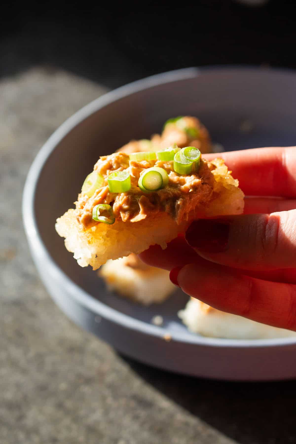 A hand holding a crispy rice bites topped with tuna and spring onion slices.
